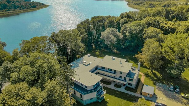 An aerial view of a modern two-story building with a flat roof, surrounded by dense greenery and a large body of water.