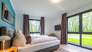 A modern bedroom with a large bed, a bedside table with a lamp, a TV mounted on the wall, and large windows offering a view of the outdoors. - 5 star vacation apartment "Waldameise" with terrace in Krakower am See (Krakow am See)