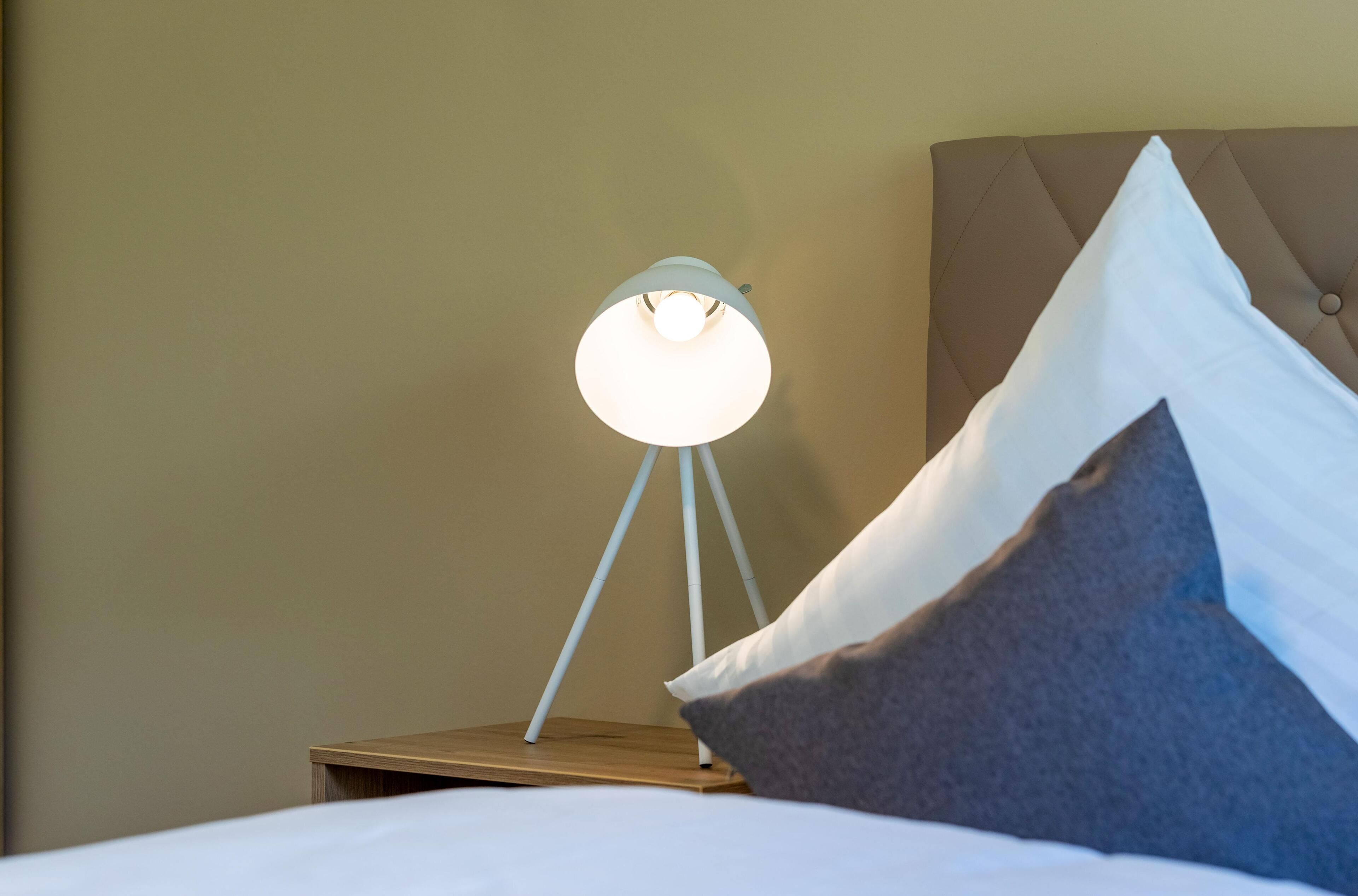 A neatly made bed with white bedding, a bedside lamp with a white shade, and a brown headboard.
