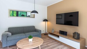 A modern living room with a grey sofa, a round wooden coffee table with a plant, a floor lamp, a TV on the wall, and a wooden cabinet with a statue. - 5 star vacation apartment "Frau Hummel" on Lake Krakow (Krakow am See)