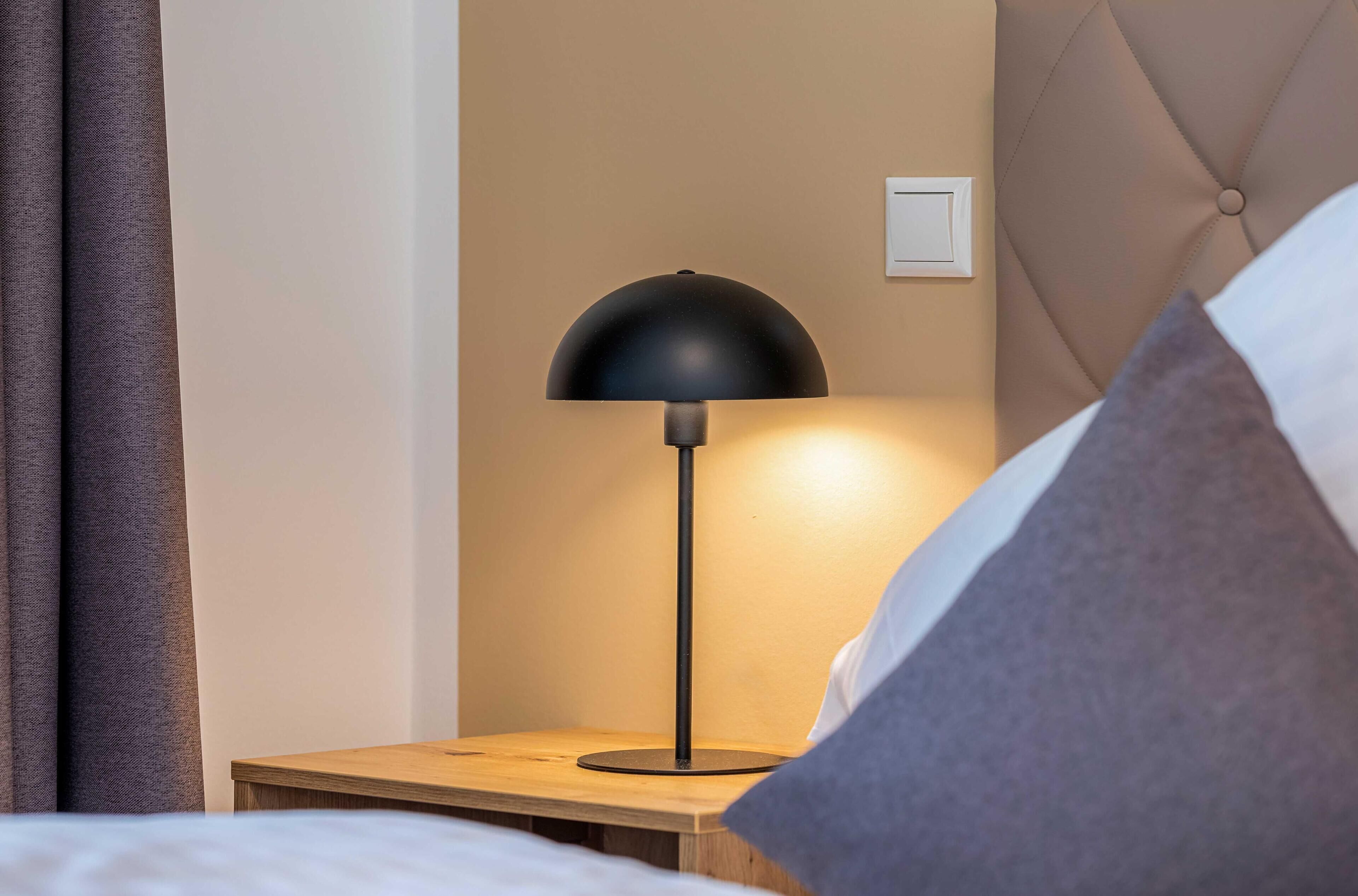 Ein ordentlich bezogenes Bett mit einem Holzkopfstück, einem Nachttisch mit Lampe und einem Lichtschalter an der Wand.