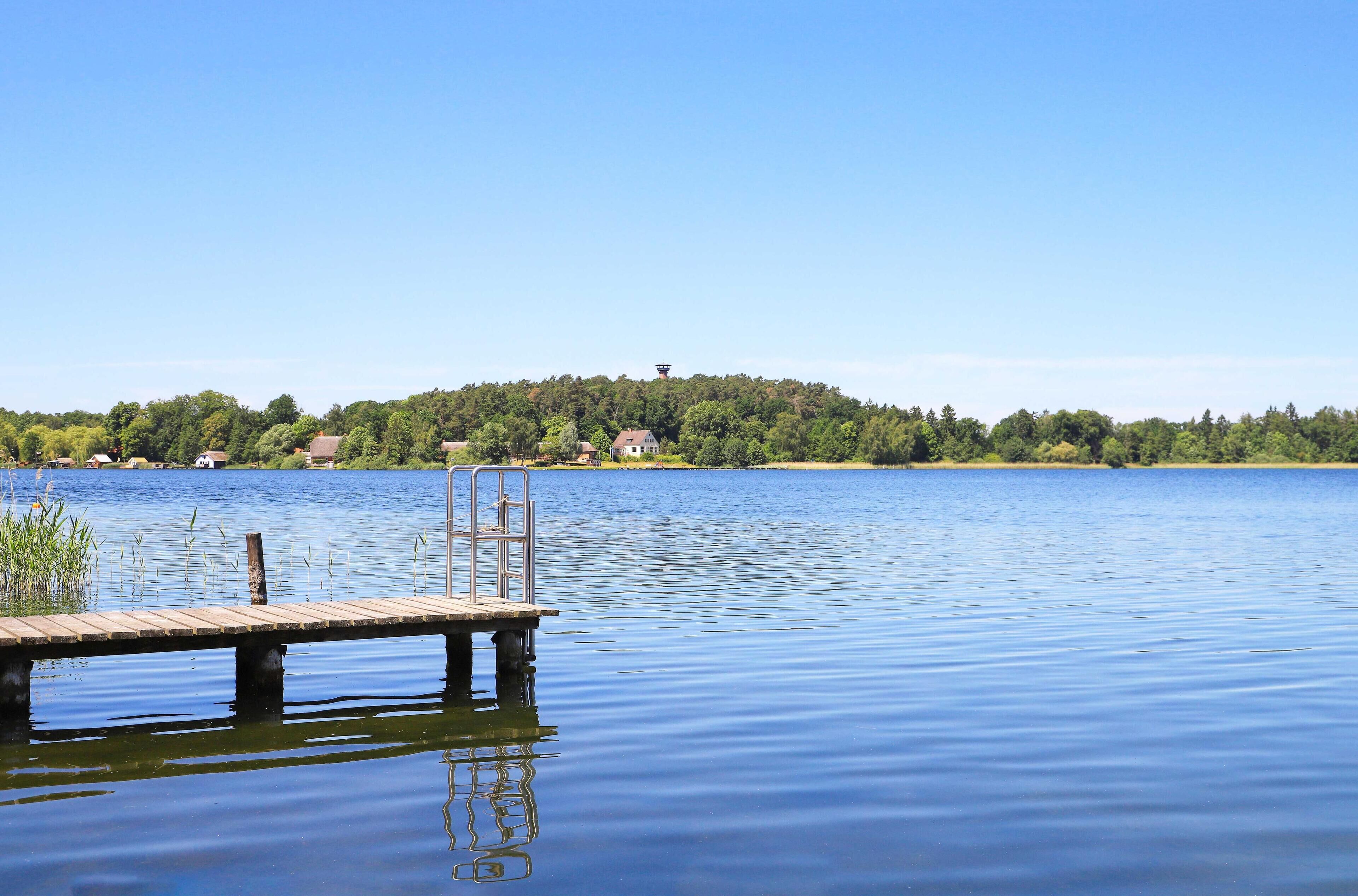 Eine hölzerne Mole führt ins ruhige Wasser, im Hintergrund ist ein bewaldeter Hügel zu sehen.