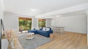 A spacious living room with a blue sectional sofa, a round coffee table, and a dining table with chairs. Large windows provide natural light and a view of greenery outside.