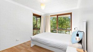 A bedroom with a bed, two windows, a bedside lamp, and a view of trees.