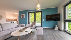 En moderne stue med en grå sofa, et rundt sofabord, en grå lænestol og en meditationsfigur. Et fjernsyn er monteret på væggen, og et stort vindue giver udsigt til grøn vegetation udenfor.
