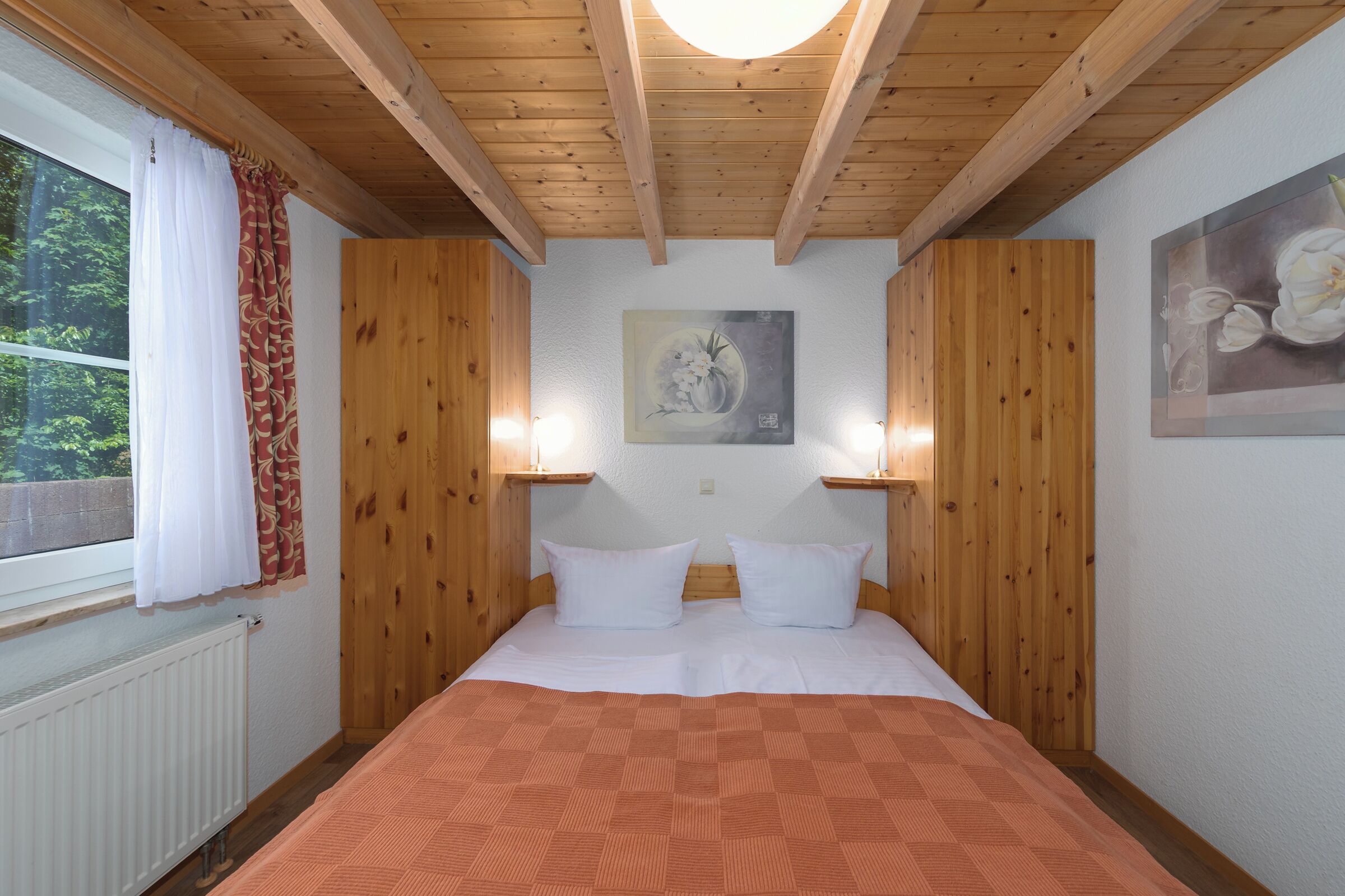 Une chambre avec des lambris en bois, un lit recouvert de draps blancs, une fenêtre avec des rideaux et des luminaires fixés au mur.