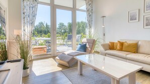 A modern living room with a sofa, a coffee table, and a chair. There are large windows with curtains, a lamp, and a plant.