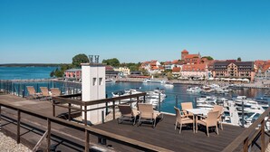 Una terraza de madera con sillones y mesas da vista a una marina con barcos y edificios al fondo.