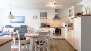 A modern kitchen with white cabinets, a dining area with a wooden table and white chairs, and a living room with a blue sofa and a painting of sailboats.