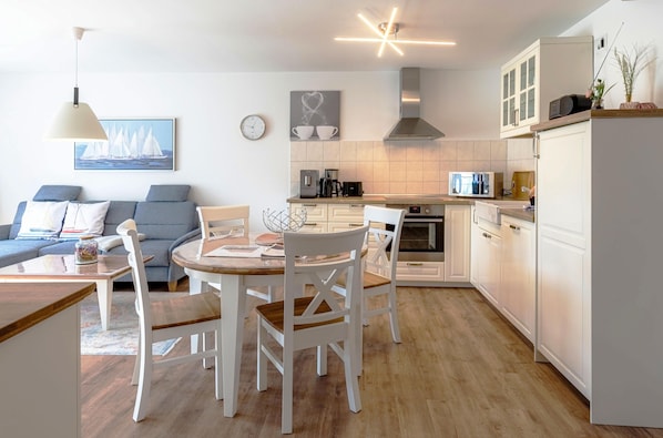 A modern kitchen with white cabinets, a dining area with a wooden table and white chairs, and a living room with a blue sofa and a painting of sailboats.