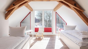 A bedroom with a sloped ceiling, wooden beams, a large window, a chair, and two beds.