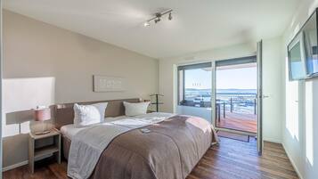 A modern hotel room with a large bed, a bedside table with a lamp, a TV mounted on the wall, and a sliding glass door leading to an outdoor area with a view of the sea.