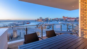 Un balcon doté de tables et de chaises en bois, offrant une vue sur une marina avec des bateaux et des bâtiments.