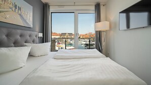 A hotel room with a large bed, a flat-screen TV, a city view through the windows, and a chandelier. - Vacation apartment with water view on the 1st floor of the Hafenresidenz Waren (Waren (Müritz))
