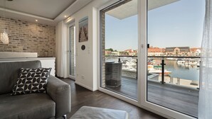 A modern living room with a grey sofa, a patterned pillow, and a view of a marina through large sliding glass doors. - Vacation apartment with water view on the 1st floor of the Hafenresidenz Waren (Waren (Müritz))