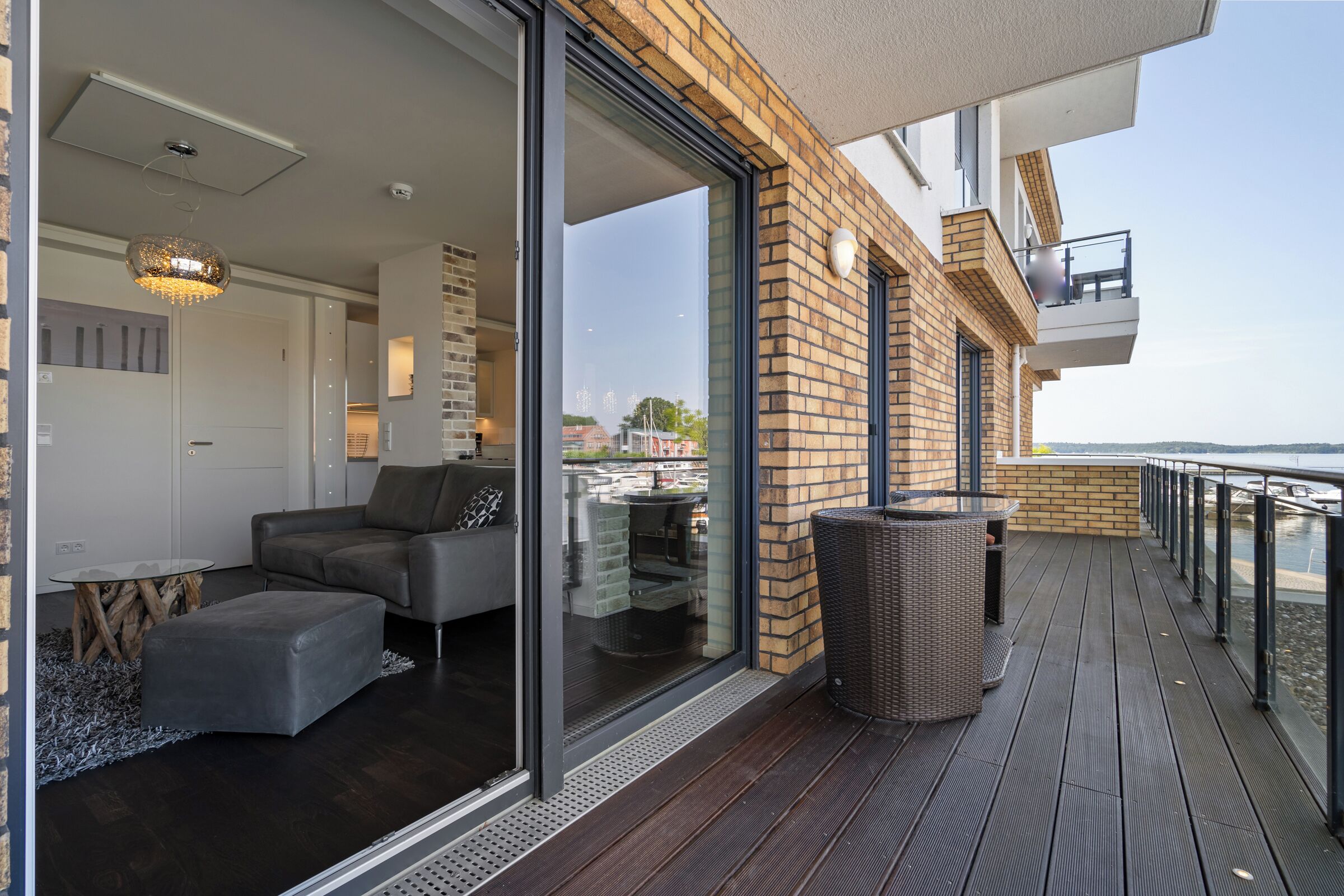 A modern living area with a sofa, ottoman, and glass doors leading to a balcony with a wicker chair and a trash bin.