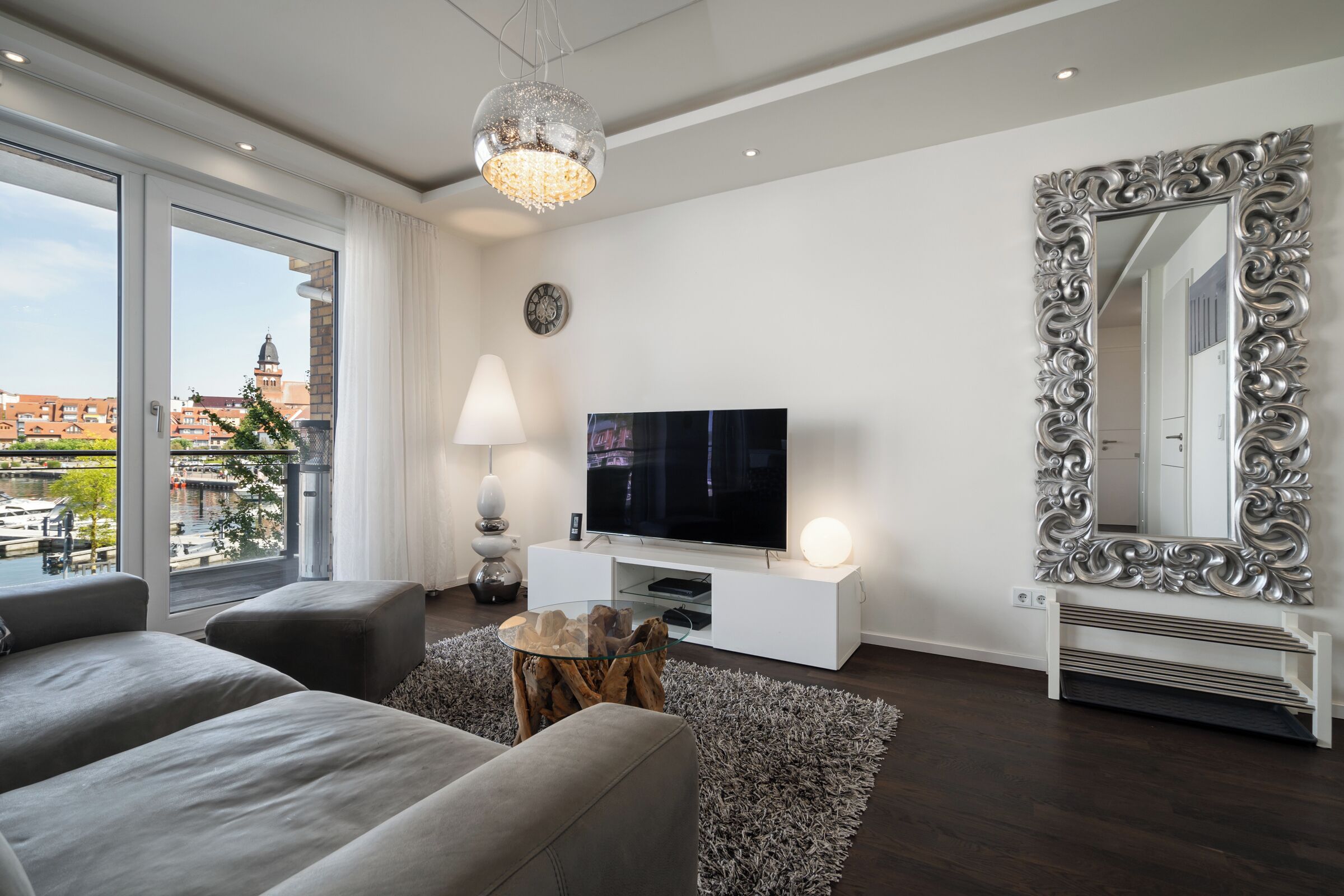 A modern living room with a large TV, a decorative mirror, and a view of the city through large windows.