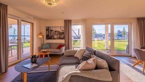 En moderne stue med sofa, sofabord og en lysekrone. Store vinduer med gardiner og udsigt til det fri.