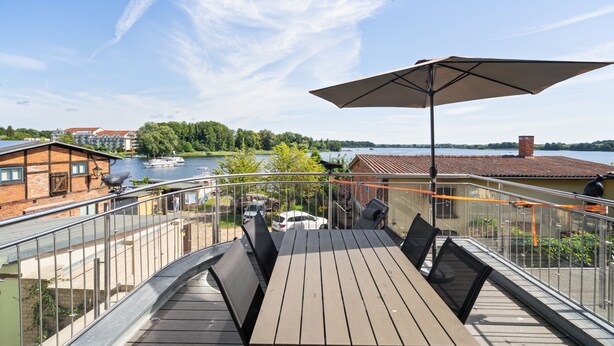A balcony with a wooden table and chairs, overlooking a body of water and buildings.