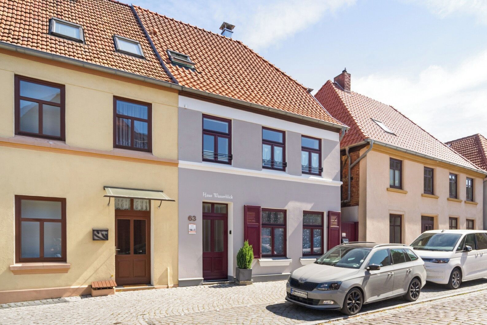 Une rangée de bâtiments de style européen traditionnel, une rue pavée, une enseigne indiquant « Haus Wasserblick » et deux voitures garées.