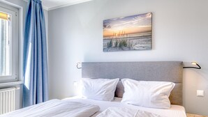 Un lit bien fait, avec du linge de lit blanc, une tête de lit grise et un tableau représentant une scène de plage accroché au mur.