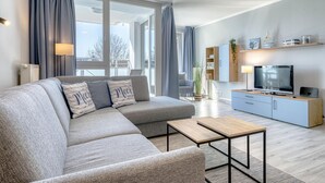 Un salon moderne avec un canapé d’angle gris, une table basse en bois, un téléviseur posé sur un meuble en bois et un balcon doté de grandes fenêtres.
