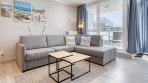 Un salon moderne avec un canapé gris, une table basse en bois et une vue sur l’extérieur grâce à de grandes fenêtres.