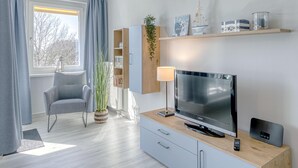 Un salon moderne avec un téléviseur, un fauteuil, une lampe et une étagère ornée d’objets décoratifs.