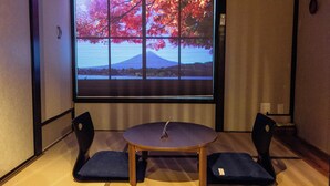 Una stanza con pavimento in tatami, un tavolino in legno, due poltrone imbottite blu e un'ampia finestra che offre una vista panoramica su una montagna e il foliage autunnale.