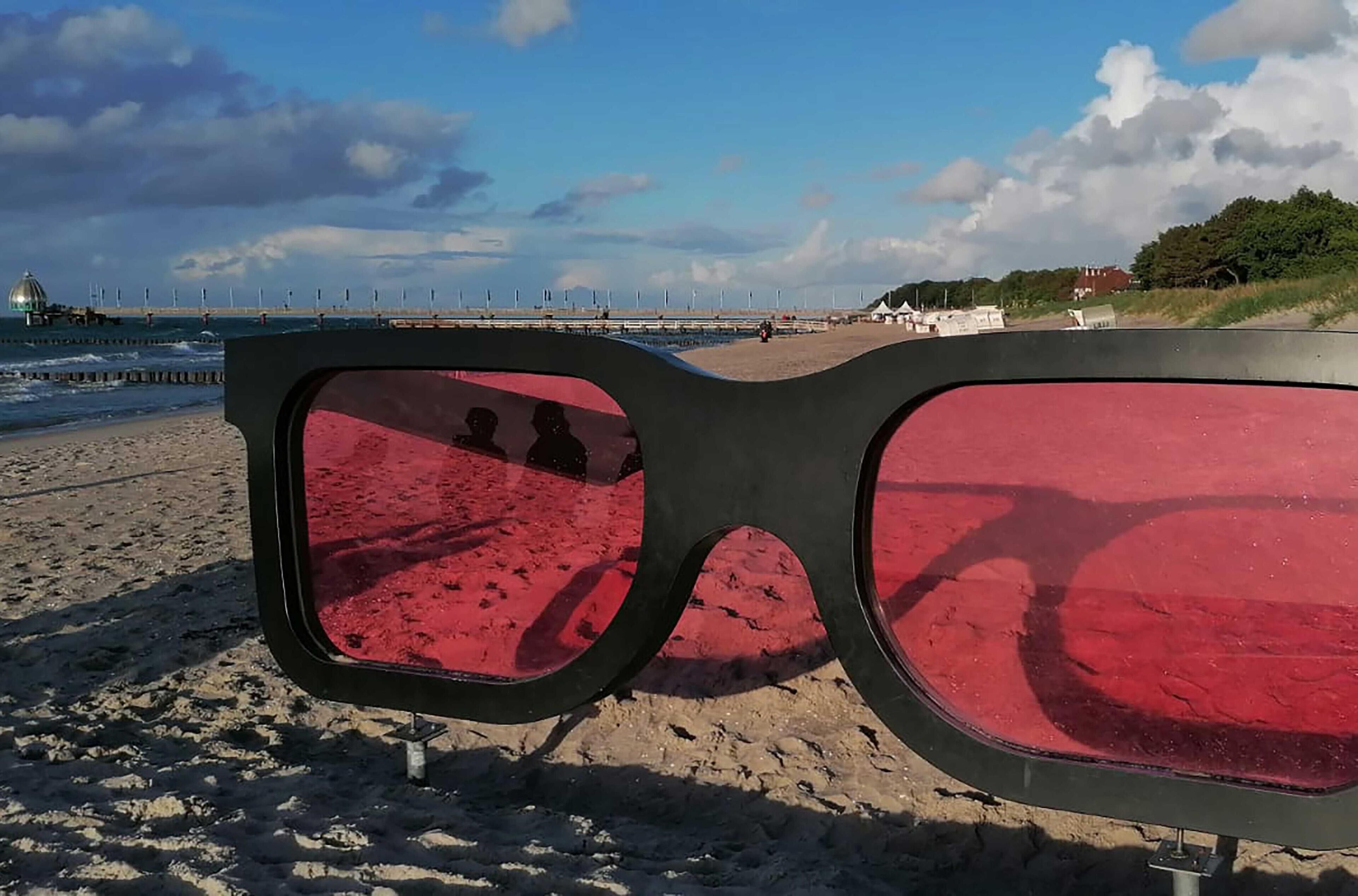 A pair of sunglasses on a sandy beach with a view of a pier and buildings in the background.