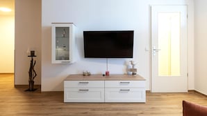 Un salon moderne avec un téléviseur à écran plat fixé au mur, un meuble en bois avec des tiroirs, un meuble en verre et une statue d’oiseau décorative.