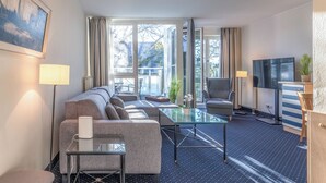 A modern hotel room with a sofa, a glass coffee table, a TV, and a desk.