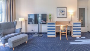 A hotel room with a sofa, a desk with drawers, a television, a lamp, a chair, and a small plant.