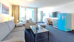 Une chambre d’hôtel avec un canapé, une table basse en verre, un réfrigérateur bleu, une télévision et un coin repas comprenant une chaise et une table.