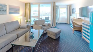 Une chambre d’hôtel comprenant un canapé, un fauteuil, un pouf, une table basse en verre et un bureau avec une chaise. Il y a un téléviseur accroché au mur et une fenêtre avec des rideaux.