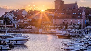 Un port de plaisance avec des bateaux amarrés, un bâtiment imposant surmonté d’une tour, et un coucher de soleil en arrière-plan.