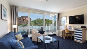 A modern hotel room with a blue sofa, a glass coffee table, a desk with a computer, a TV, and a balcony with a view of the sea.