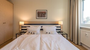 A hotel room with a large bed, two bedside tables, a headboard with a picture, and a window with curtains. - "Kranichtanz" Vacation Rental with a Balcony and a View Toward the Dike (Zingst)