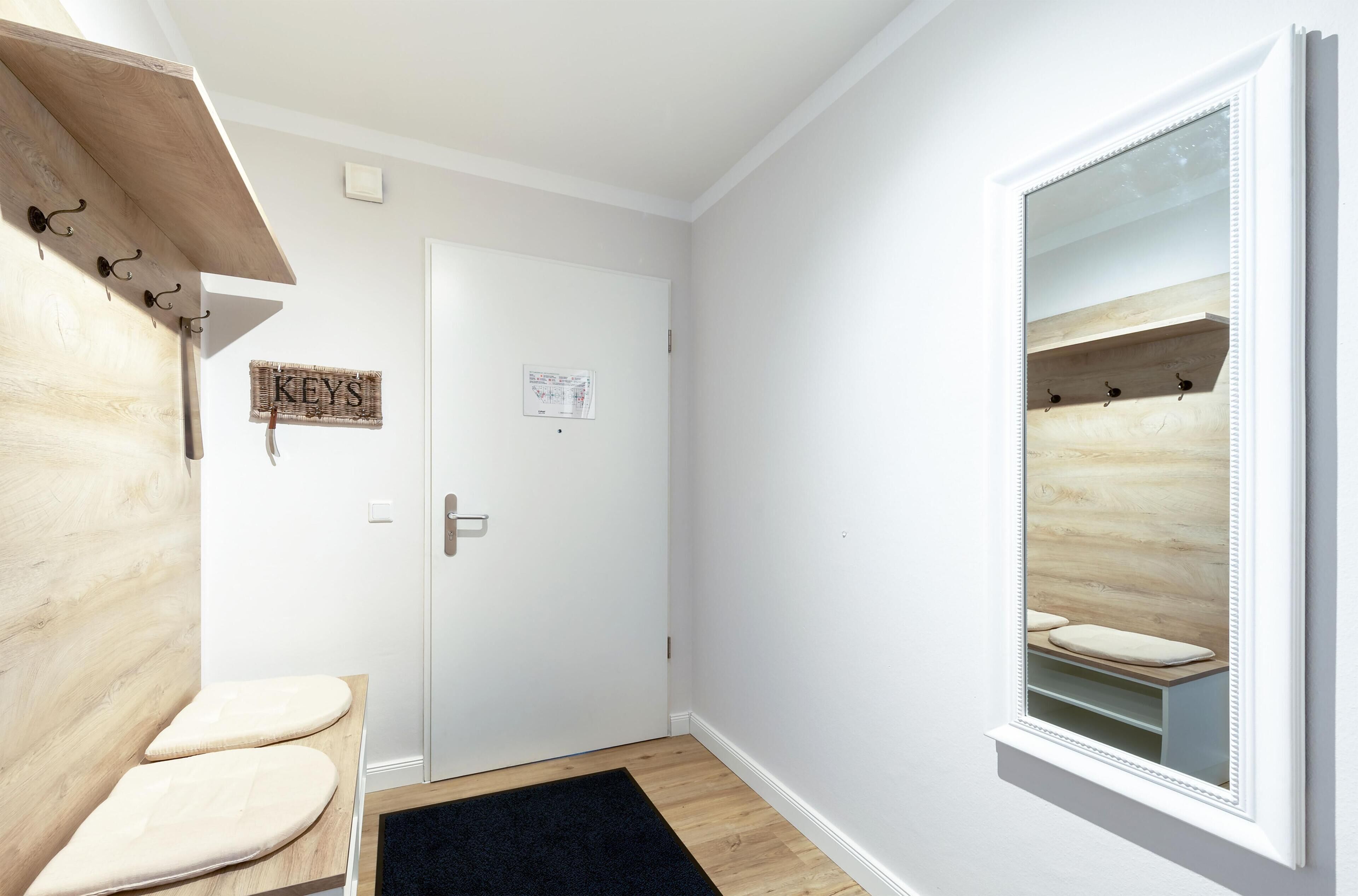 Un couloir avec un banc en bois, une porte sur laquelle est apposée une pancarte indiquant « CLÉS », et un miroir qui reflète une étagère en bois.