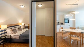 A modern hotel room with a bed, bedside table, lamp, and a view into the kitchen area.