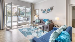 Una sala moderna con una mesa de centro de vidrio, un sofá y un sofá azul. Hay una ventana grande con cortinas, un adorno en la pared y una lámpara en una mesita auxiliar.
