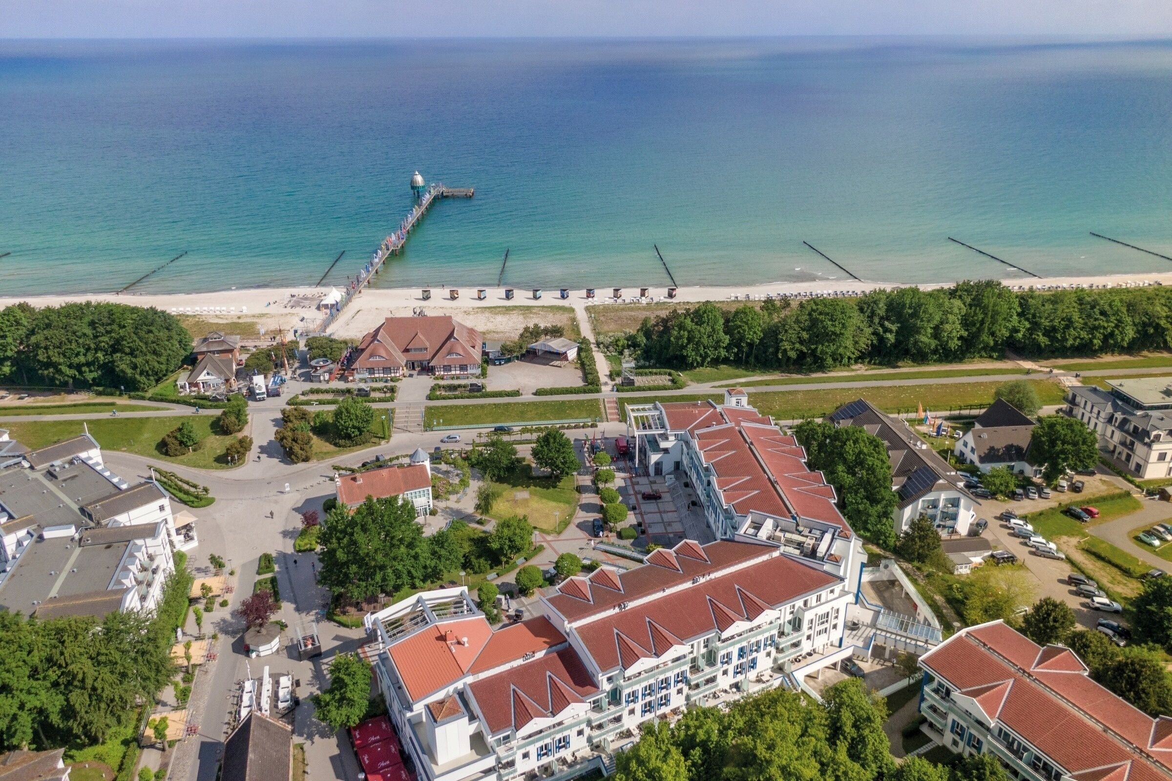 Luchtbeeld van een kusthotel met meerdere gebouwen, een steiger die in zee uitloopt, en een strand op de achtergrond.