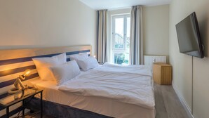 A neatly made bed with white linens, a bedside table with a lamp, a TV mounted on the wall, and a window with curtains.