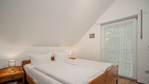 Un dormitorio con cama de madera, ropa de cama blanca, una mesita de noche con lámpara y una ventana con cortina blanca.