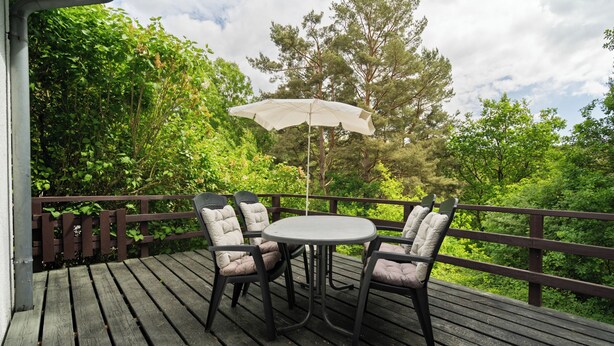 Una terraza de madera con una mesa redonda y cuatro sillas, una sombrilla, y vistas de vegetación exuberante y árboles.
