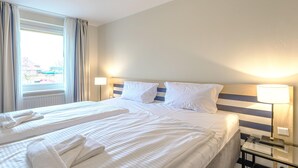 Habitación de hotel con dos camas, un ventanal con cortinas, una mesita de noche con lámpara y vistas a edificios.