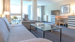 Habitación de hotel con sofá, una mesita de vidrio, televisión, escritorio y ventana con cortinas.