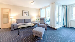 Habitación de hotel con un sofá gris, un sillón, un reposapiés, una mesa de centro de cristal y un ventanal con cortinas.