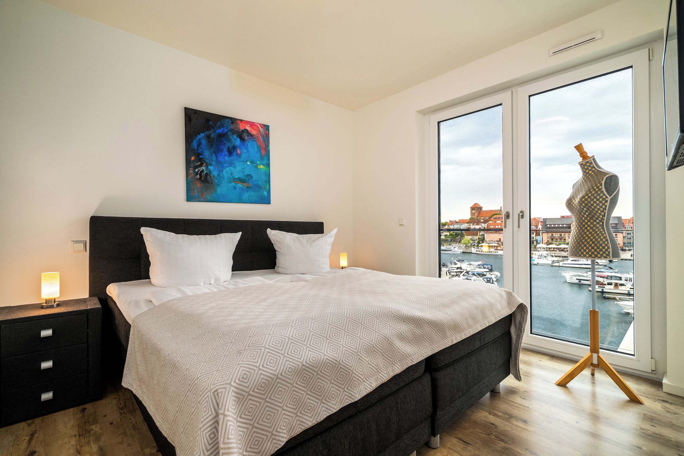 A modern bedroom with a large bed, a nightstand, a lamp, and a view of a marina through a glass door.