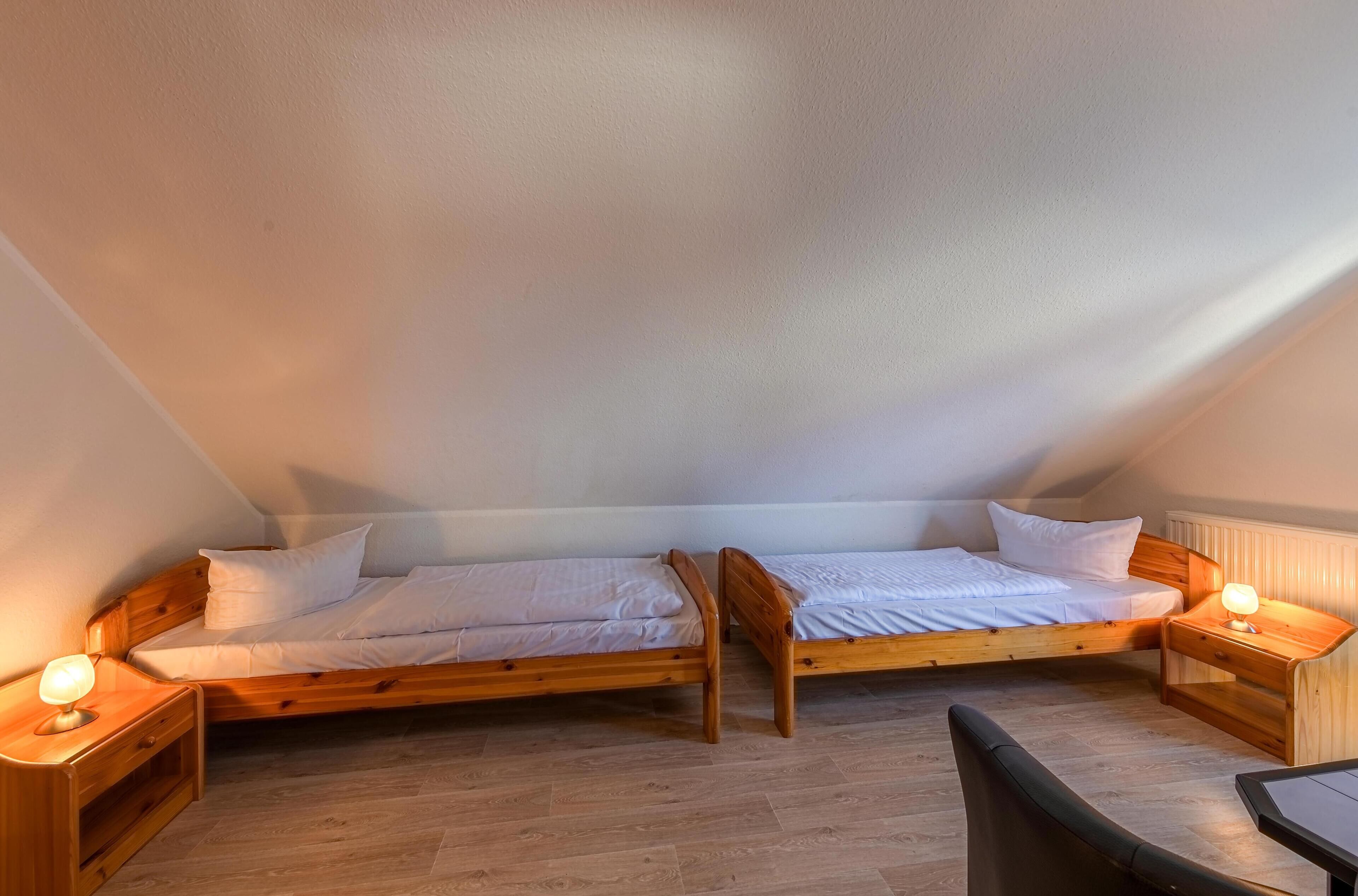 A small, cozy attic bedroom with two wooden beds, nightstands with lamps, and a wooden floor.